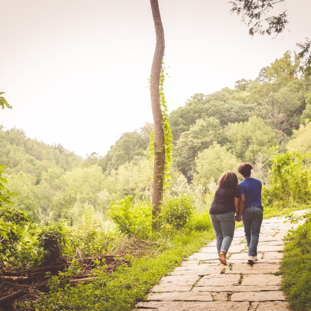 A couple walks hand in hand on a sunlit stone path through a lush green forest, conveying a sense of tranquility and togetherness.