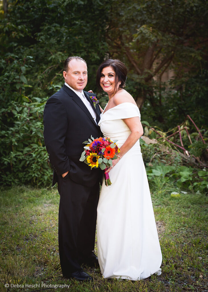 Bride and groom smiling together in front of lush greenery at their Cutillo's Restaurant wedding.