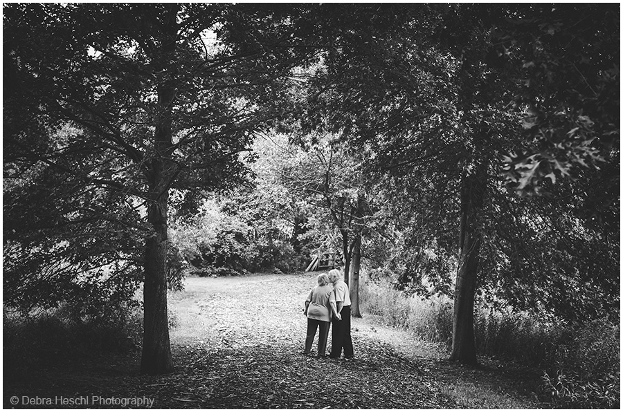 Black and white photo of an elderly couple walking hand in hand down a wooded path during their anniversary photo session.