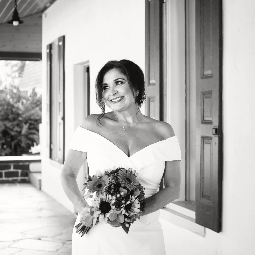 A black & white photo of a beautiful bride  standing under a porch with a bouquet of flowers.