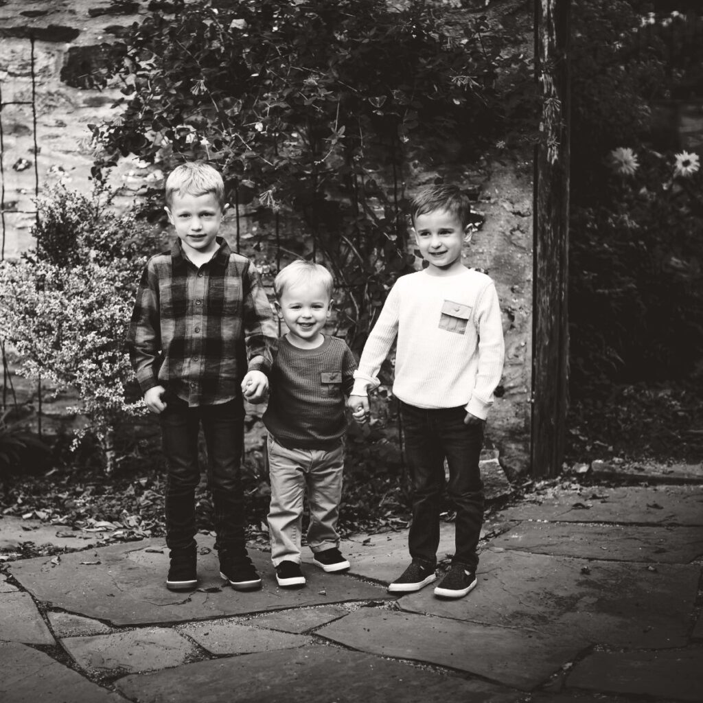 Three young boys stand holding hands on a stone path in a garden. They smile warmly, dressed in casual shirts and pants, surrounded by lush foliage. Black and white.