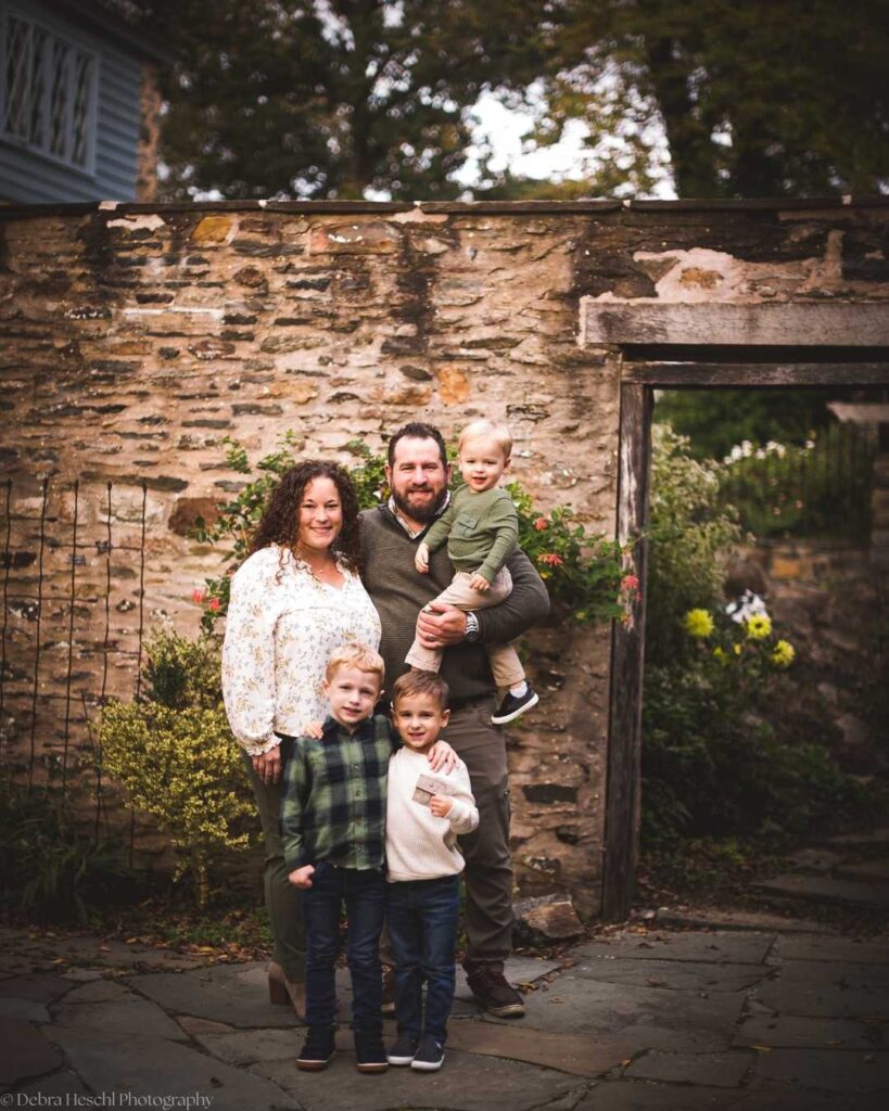 A family of five poses outdoors against a rustic stone wall. The parents smile warmly, holding their three young children. The scene is cozy and inviting.