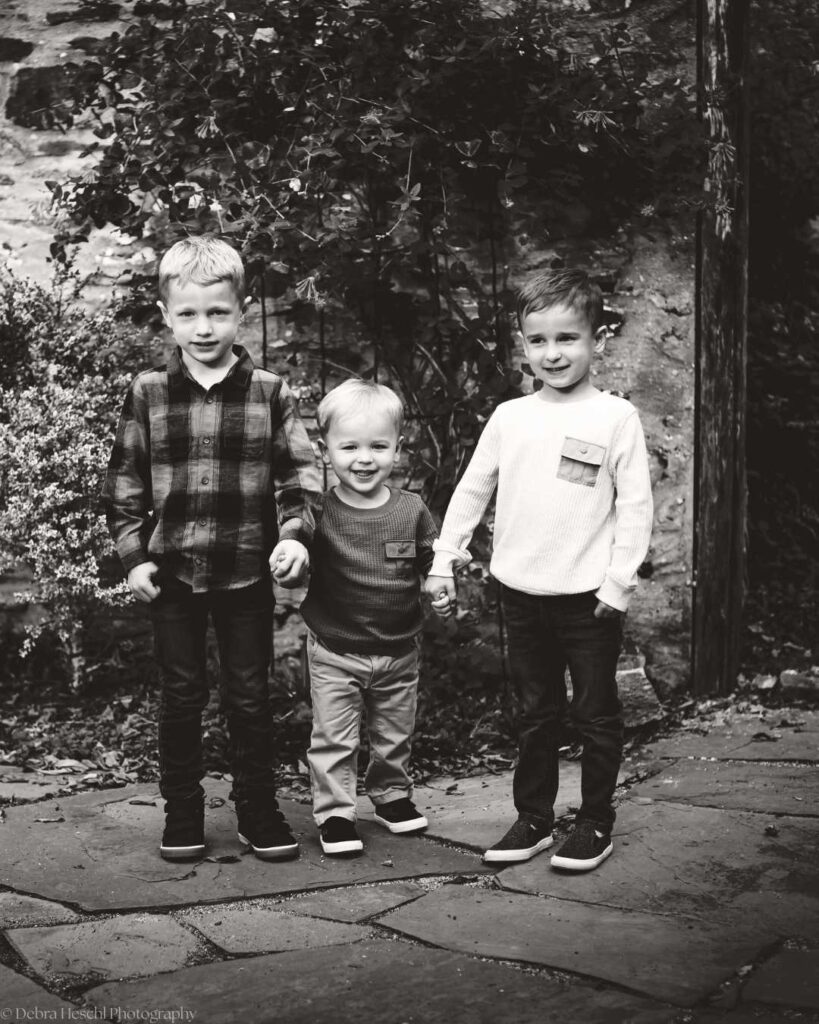 Three young boys stand holding hands on a stone path in a garden. They smile warmly, dressed in casual shirts and pants, surrounded by lush foliage. Black and white.