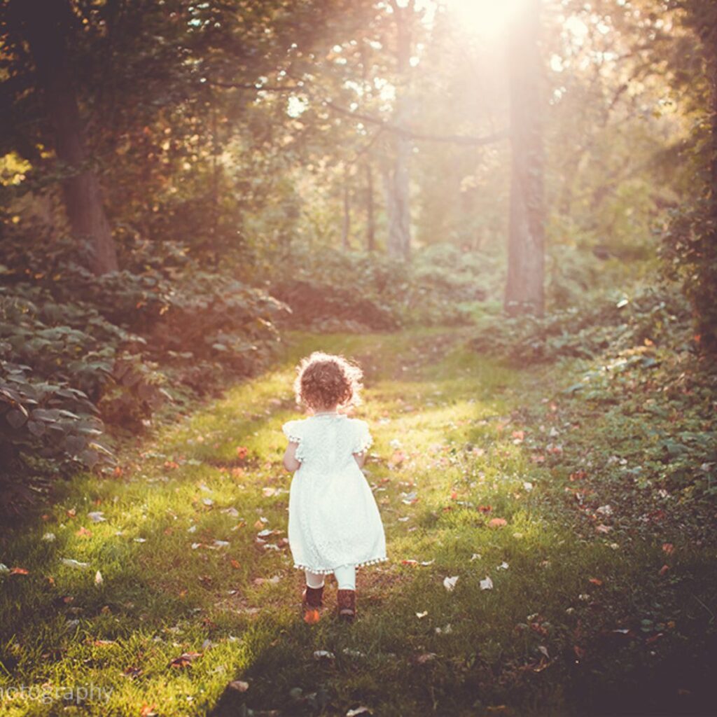 A little girl with bouncing curls in a white dress walking away in a pathway of grass with a sun rays highlighting her hair.