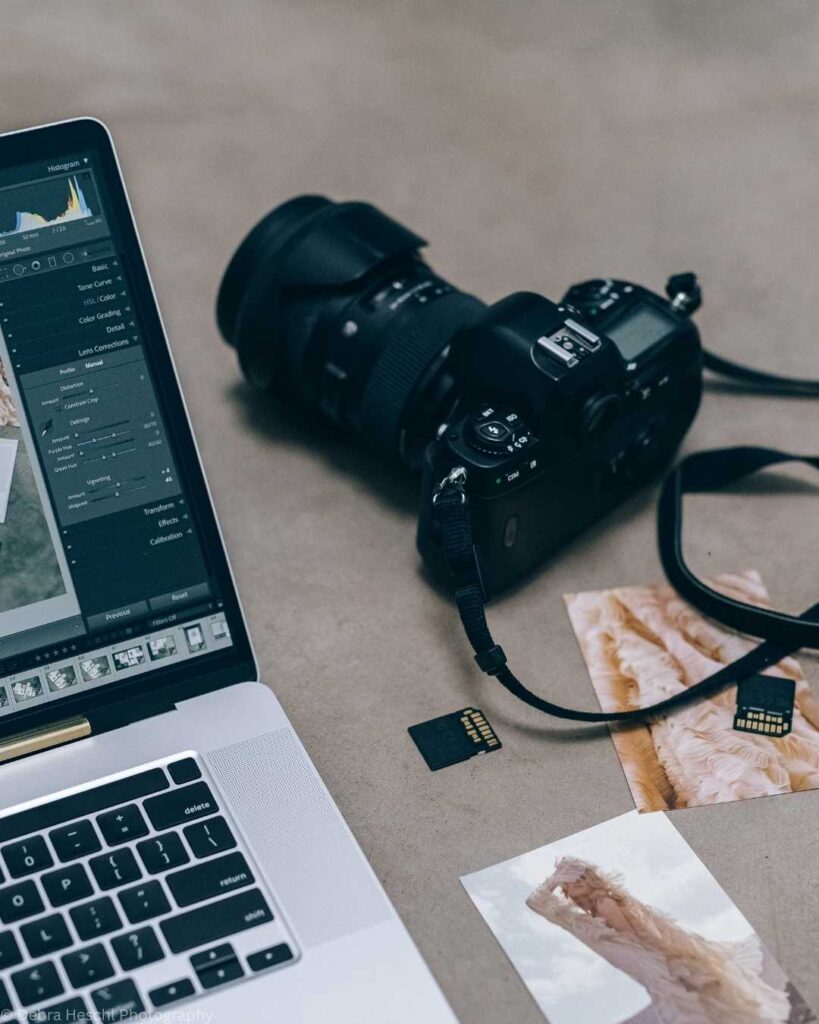 Laptop with photo editing software, DSLR camera, memory cards, and printed photos on a desk. The scene conveys a focused and creative atmosphere.