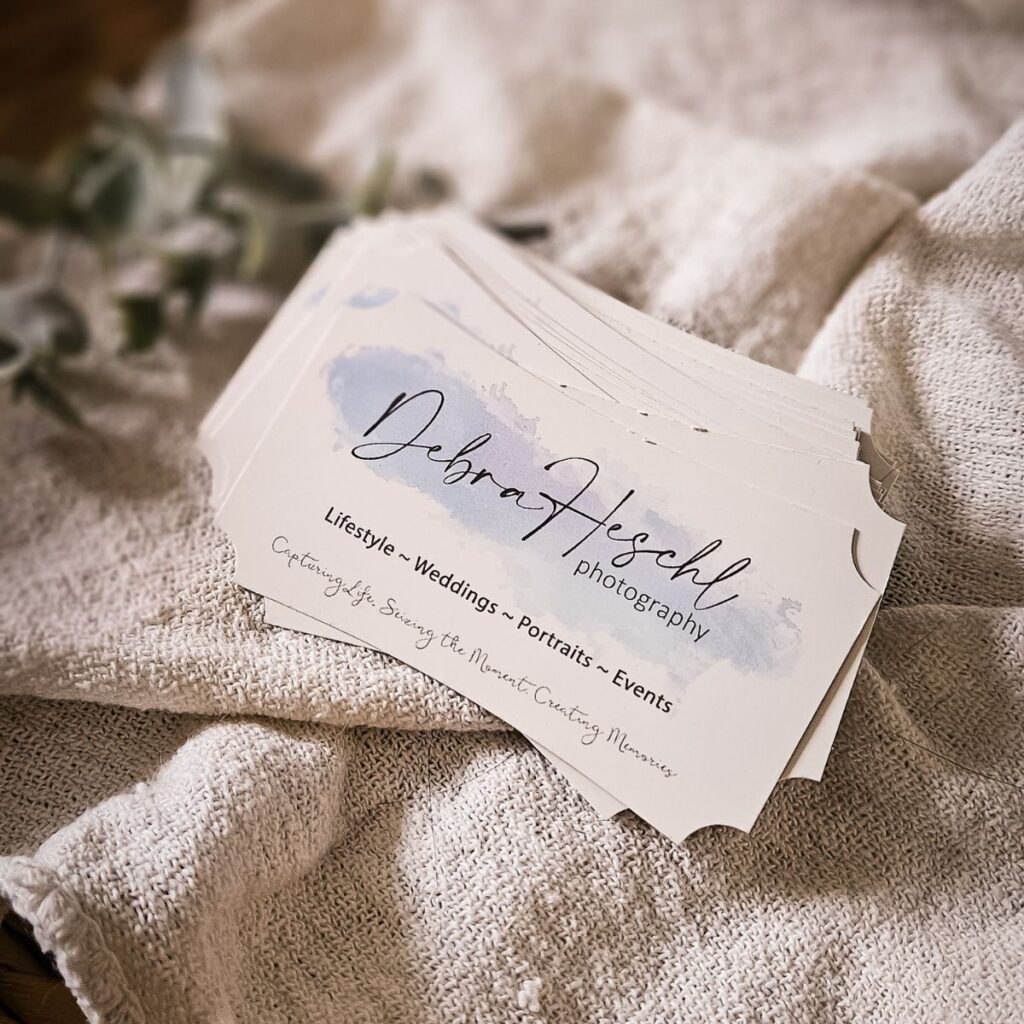 A stack of elegant photography business cards on a soft textured cloth. Cards display delicate script with "Lifestyle, Weddings, Portraits, Events."