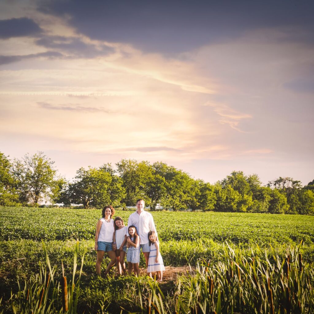 A family of five stands together in a lush green field under a colorful sunset sky. The mood is serene, and the setting evokes warmth and togetherness.