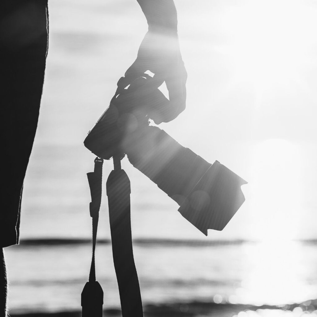 Silhouette of a person holding a camera facing the ocean with bright sunlight creating lens flares. The tone is serene and contemplative.