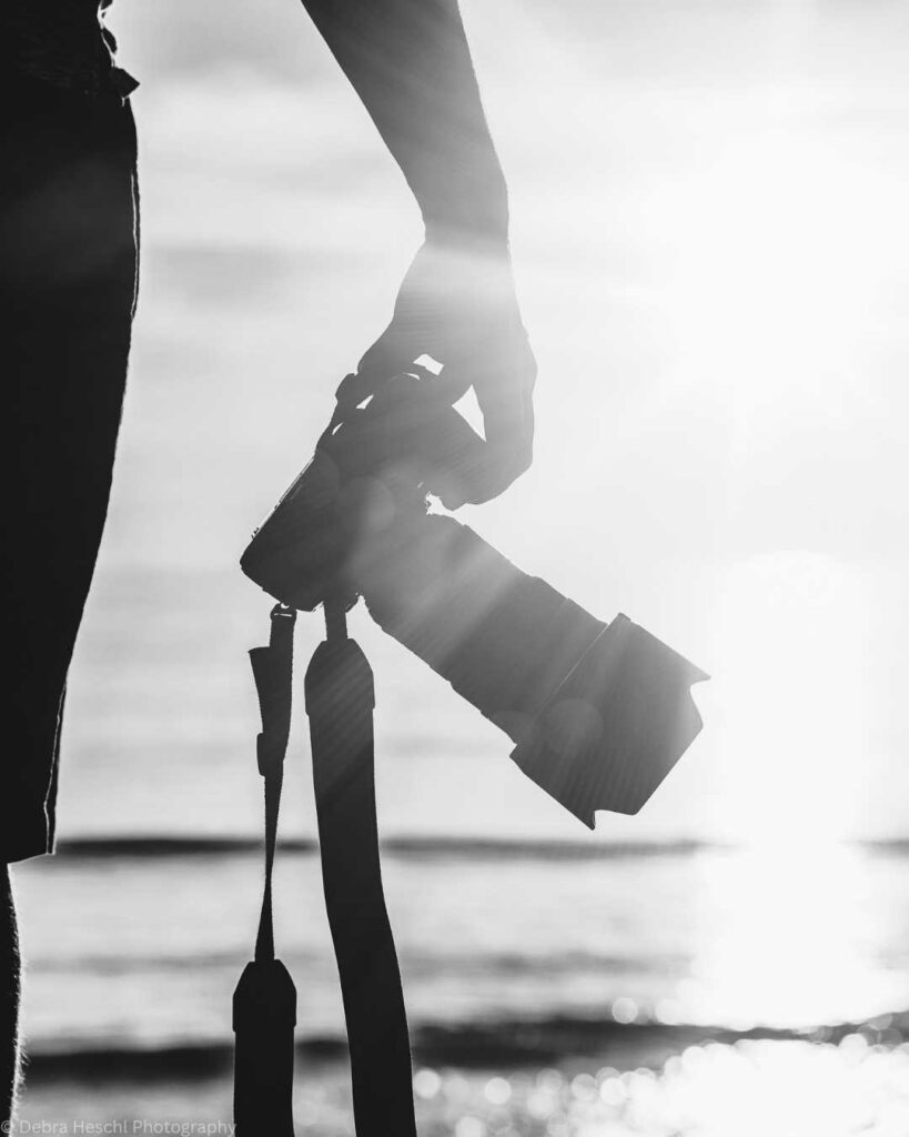 Silhouette of a person holding a camera facing the ocean with bright sunlight creating lens flares. The tone is serene and contemplative.