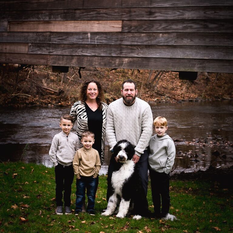 A family of five and a large black and white dog pose in front of a wooden covered bridge. They stand on green grass beside a flowing creek. The setting is calm and natural.