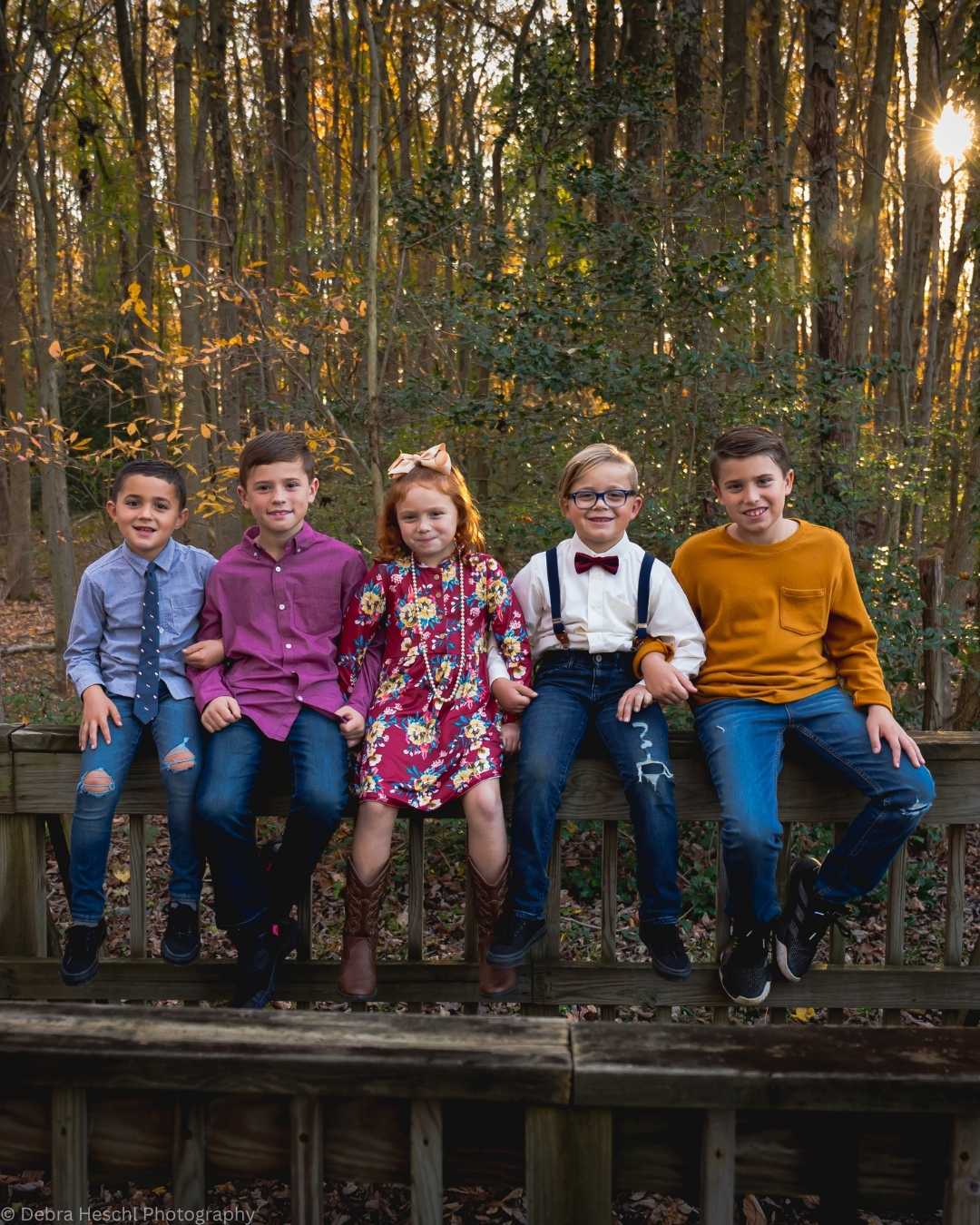 Five children sit on a wooden fence in a forest during sunset. They wear colorful outfits and smile warmly, creating a joyful, autumnal scene.