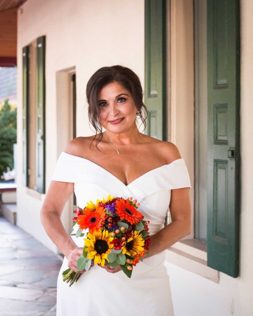A bride in an off-the-shoulder white dress holds a vibrant bouquet with sunflowers and red flowers. She smiles warmly, standing against a wall with green shutters.