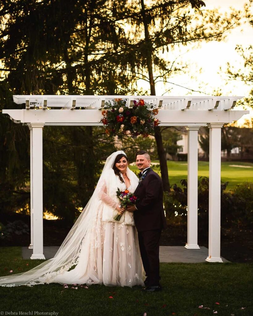 Bride and groom smiling under a white pergola adorned with flowers. The bride wears a lace dress with a flowing veil, and the groom is in a dark suit.