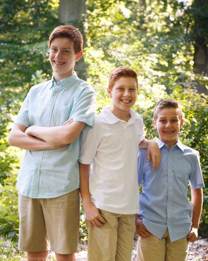 Three boys stand in a sunlit forest. The tallest is on the left, arms crossed; the middle smiles with an arm around the shortest on the right, conveying joy.