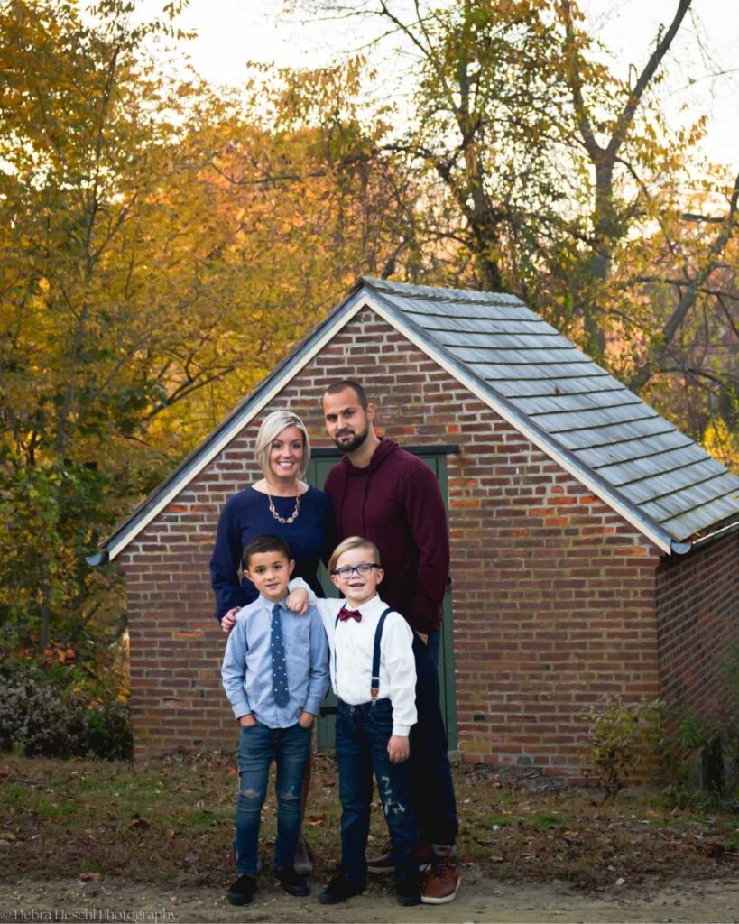 Family of four smiling in front of a small brick building surrounded by autumn trees. Parents stand behind two young boys in dressy casual attire.