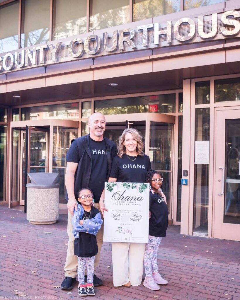 A joyful family stands in front of a courthouse. They all wear matching "OHANA" shirts. The parents hold an "Ohana" sign. Smiles convey excitement.