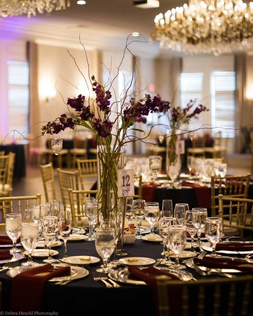 Elegant dining setup with round tables, each adorned with purple floral centerpieces and wine glasses. A chandelier hangs above, casting a warm glow.
