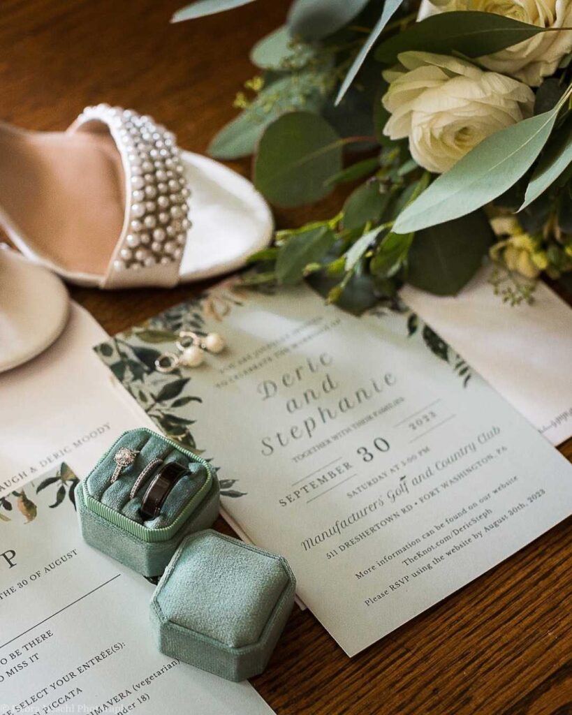 Wedding details with invitations, a pearl-embellished shoe, a floral bouquet with white roses, and mint-green ring boxes containing rings. Elegant and romantic setting.