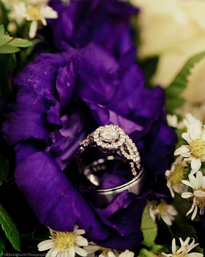 A sparkling diamond ring set rests elegantly inside a deep purple flower surrounded by delicate white daisies and green leaves, evoking romance.