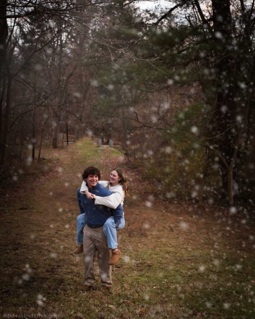 A young man gives a piggyback ride to a smiling woman on a forest path. Snowflakes fall gently around them, creating a playful and joyful atmosphere.