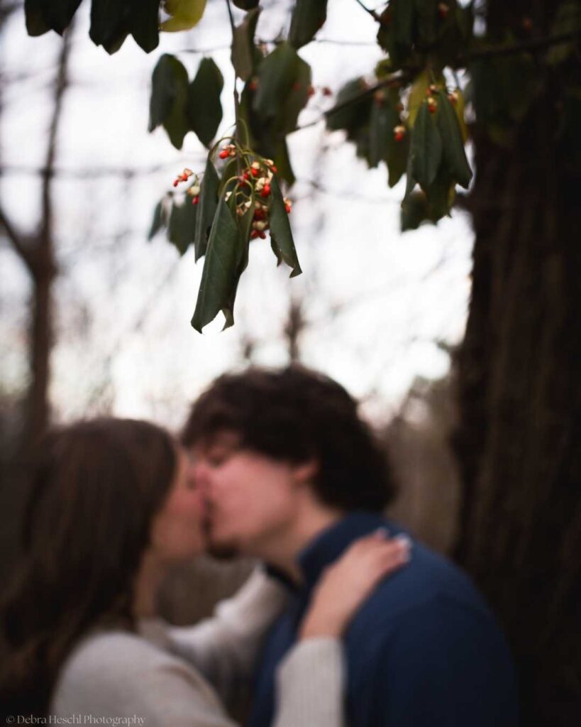 A couple shares a kiss in a blurry forest setting under a branch with green leaves and red berries, conveying a romantic and intimate mood.