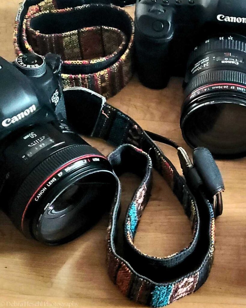 Two Canon cameras with colorful woven straps on a wooden surface. The lenses are prominently displayed, highlighting photography gear essentials.