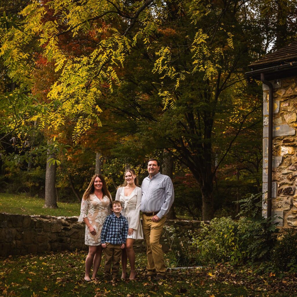 A family of four stands in front of vibrant autumn trees, with leaves in shades of yellow and orange. A stone building is visible on the right, evoking warmth.