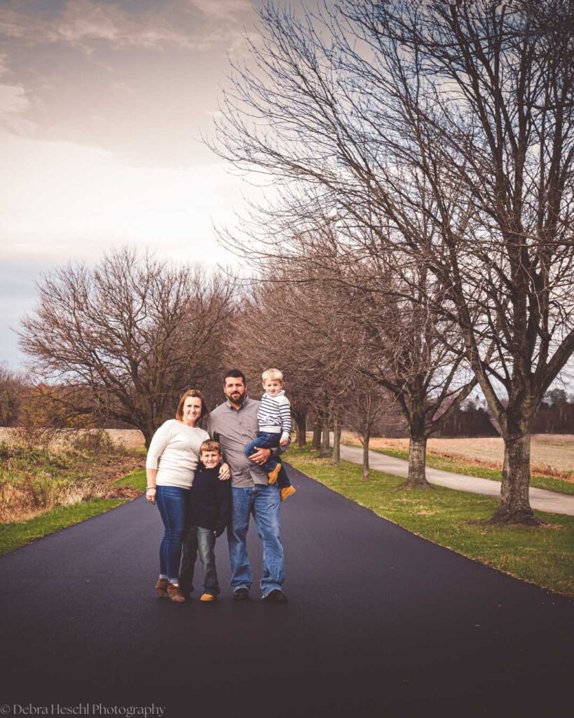 Family of four stands on a paved path lined with bare trees, under a cloudy sky. Parents and two children pose warmly, conveying a serene, autumn vibe.