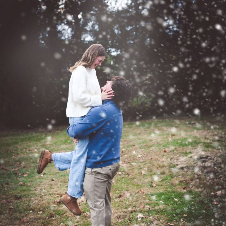 A couple embraces in a snowy outdoor scene. The woman, in a white sweater and jeans, smiles as the man, in a blue sweater, lifts her. The mood is joyful and romantic.