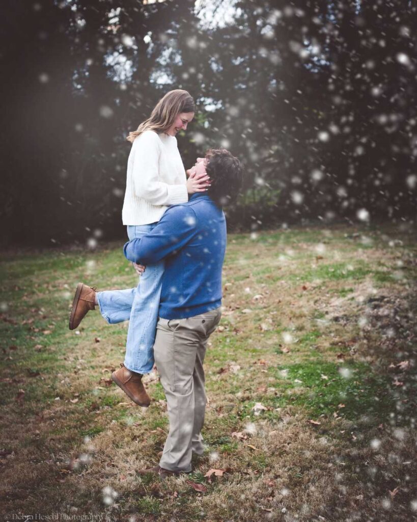 A couple embraces in a snowy outdoor scene. The woman, in a white sweater and jeans, smiles as the man, in a blue sweater, lifts her. The mood is joyful and romantic.