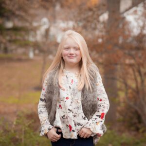 Young girl with long blonde hair stands outdoors in fall. She wears a floral blouse and gray fur vest, surrounded by autumn leaves and soft light.