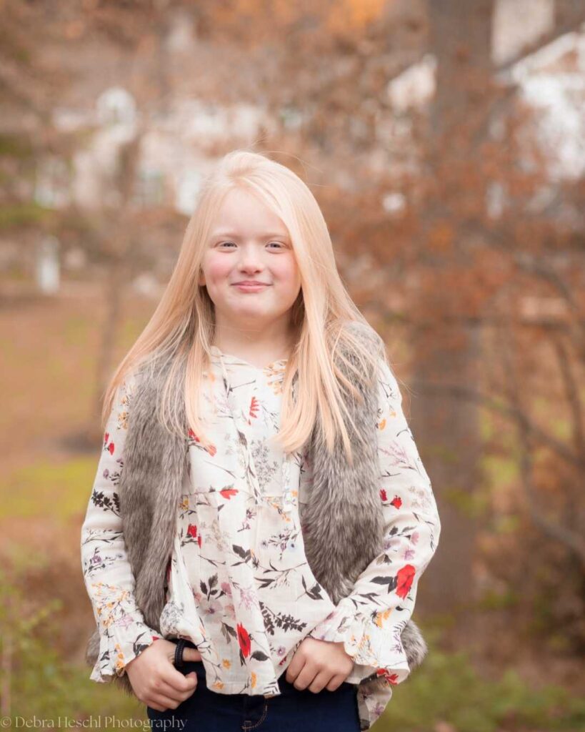 Young girl with long, blonde hair stands outdoors, wearing a floral blouse and fur vest. Autumn leaves blur in the background; her expression is serene.