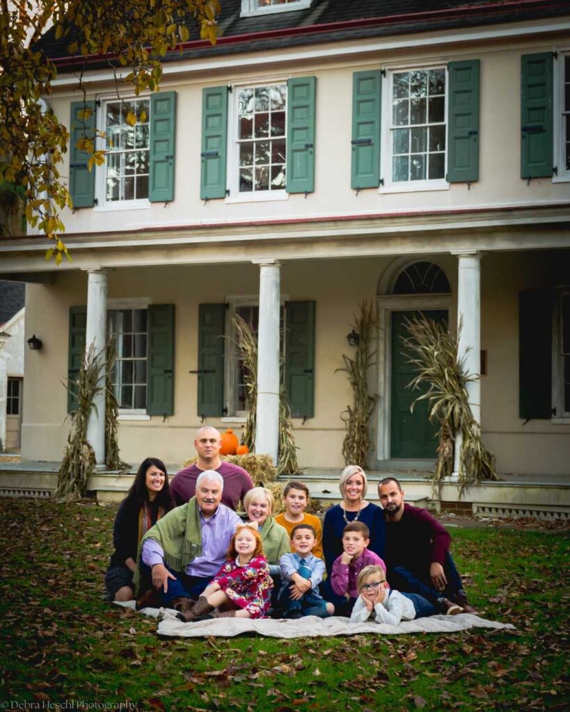 A family of twelve poses on the lawn in front of a large house with green shutters. They are sitting on a blanket, surrounded by autumn leaves, exuding warmth and happiness. Cornstalks and pumpkins adorn the porch, adding a festive, seasonal touch.