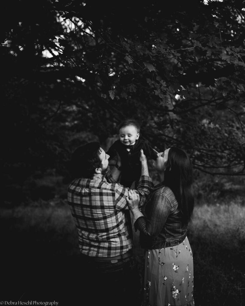 A black and white photo of a joyful family outdoors. A man in a plaid shirt lifts a smiling baby, while a woman in a floral dress watches lovingly. Trees in the background.