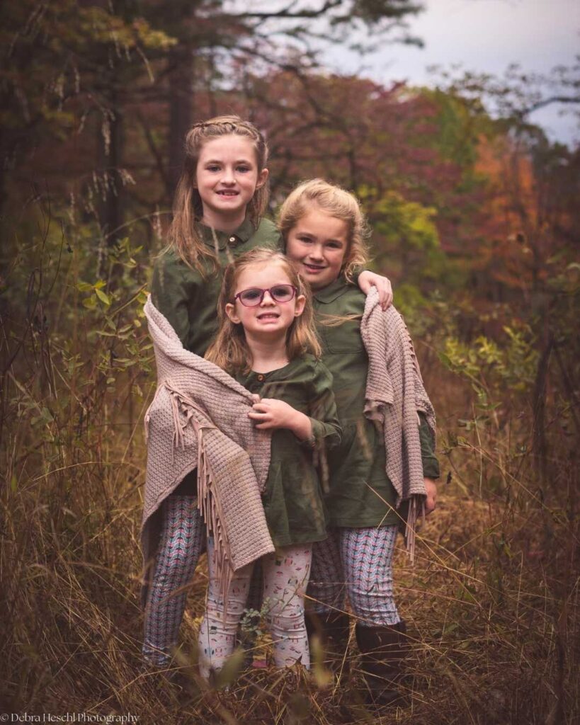 Three young girls stand closely in a grassy, autumn forest. They wear green shirts and patterned leggings, smiling warmly, wrapped in cozy shawls, conveying joy.