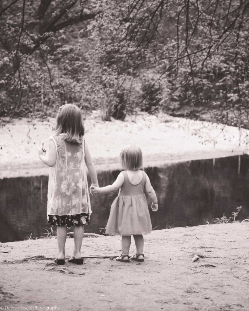 Two young girls in dresses stand hand in hand by a serene pond, surrounded by overhanging branches. The scene is calm and timeless.