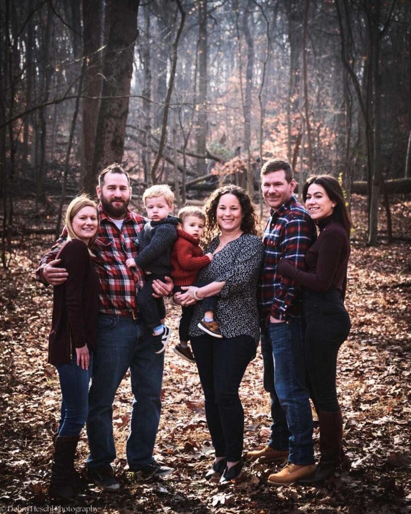 A group of six adults and two children stand together in a forest with autumn leaves. They smile warmly, dressed in casual attire, conveying a cozy, familial mood.