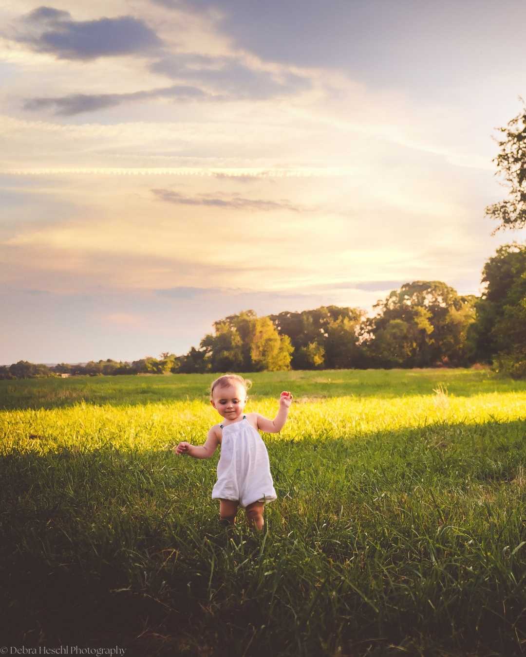 A joyful baby in a white outfit walks on a sunlit grassy field, with a backdrop of trees and a sunset sky, evoking warmth and happiness