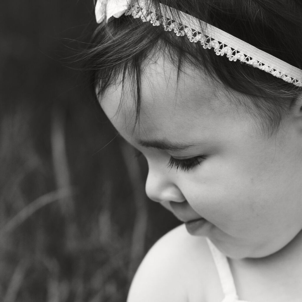 Black-and-white photo of a toddler in profile wearing a lace headband, gazing downward with a gentle, serene expression. Blurred background.