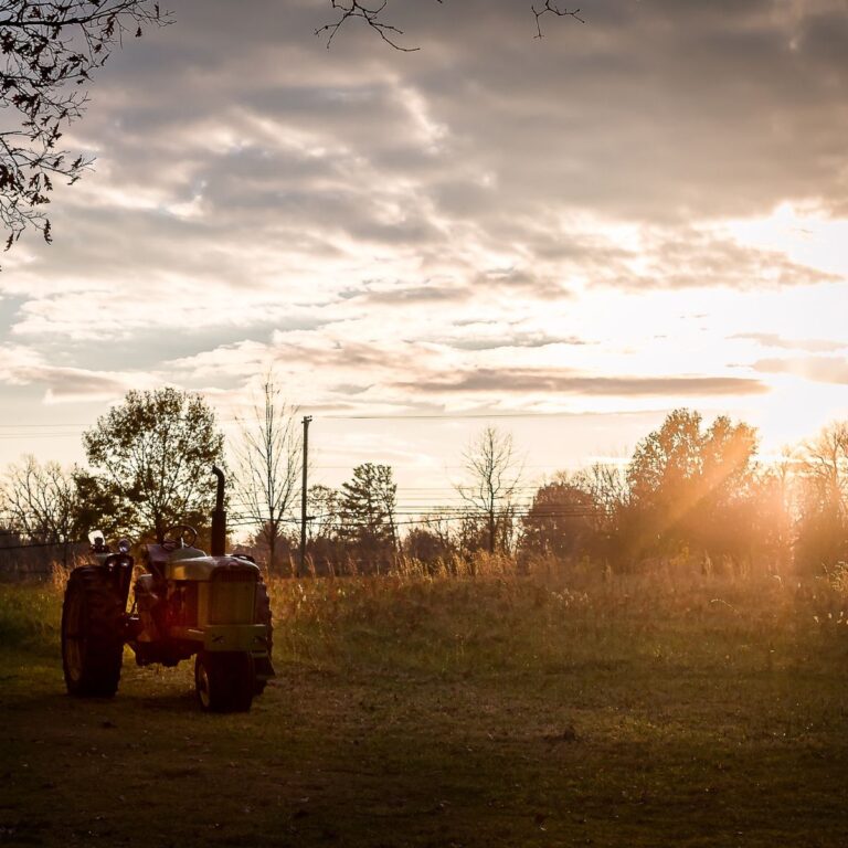 A tractor sits quietly in a sunlit field at sunset, with a warm glow and scattered clouds overhead, creating a serene rural atmosphere.