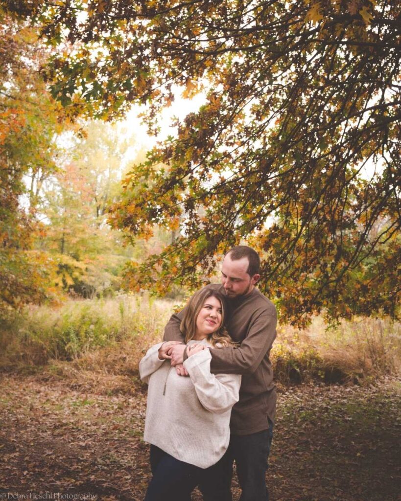 A couple embraces under a tree with autumn leaves. The woman smiles softly while the man holds her tenderly. The scene exudes warmth and intimacy.