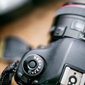 Close-up of a DSLR camera's top, showing the mode dial with various settings like auto and manual. A lens with "50mm" marking is visible, placed on a wooden surface.