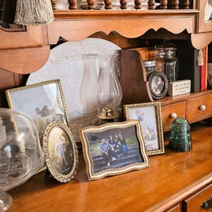 A wooden shelf displays framed family photos, an oil lamp, decorative glass items, and a Bible verse sign. The scene feels warm and nostalgic.