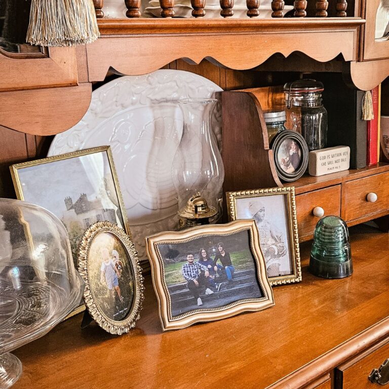 A wooden shelf displays framed family photos, an oil lamp, decorative glass items, and a Bible verse sign. The scene feels warm and nostalgic.