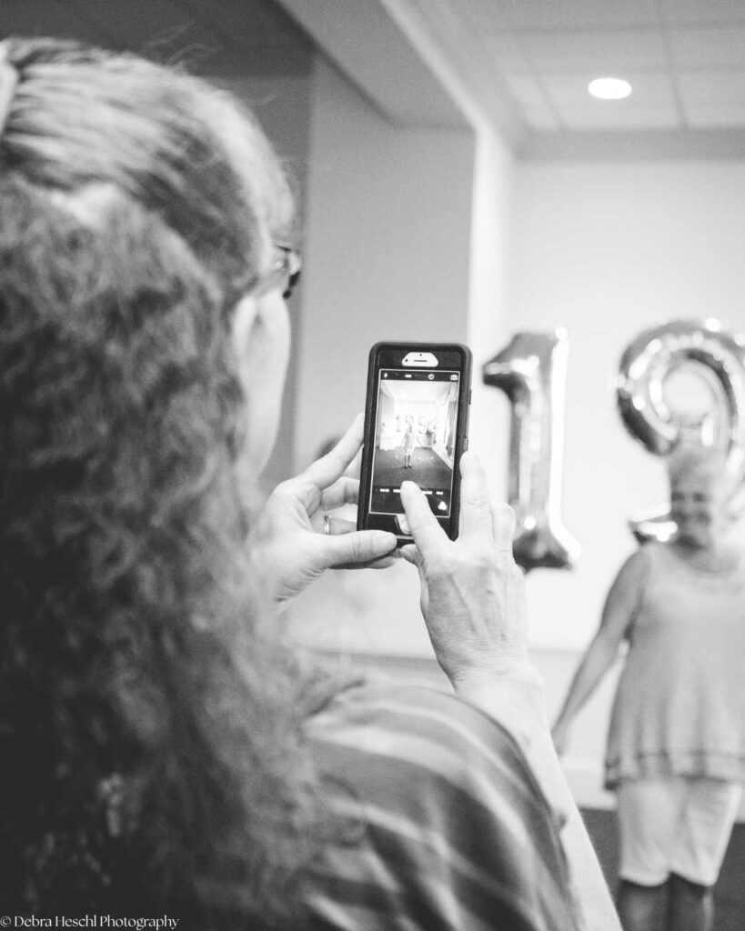 A woman with curly hair takes a photo on her phone of another smiling woman near large "19" balloons, capturing a joyful moment. Black and white.