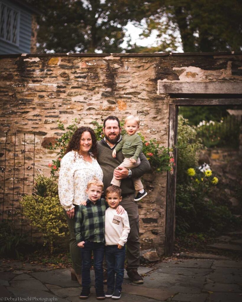 A family of five poses in front of a rustic stone wall. The parents stand behind their three young sons, one of whom is being held. The scene is warm and cheerful.