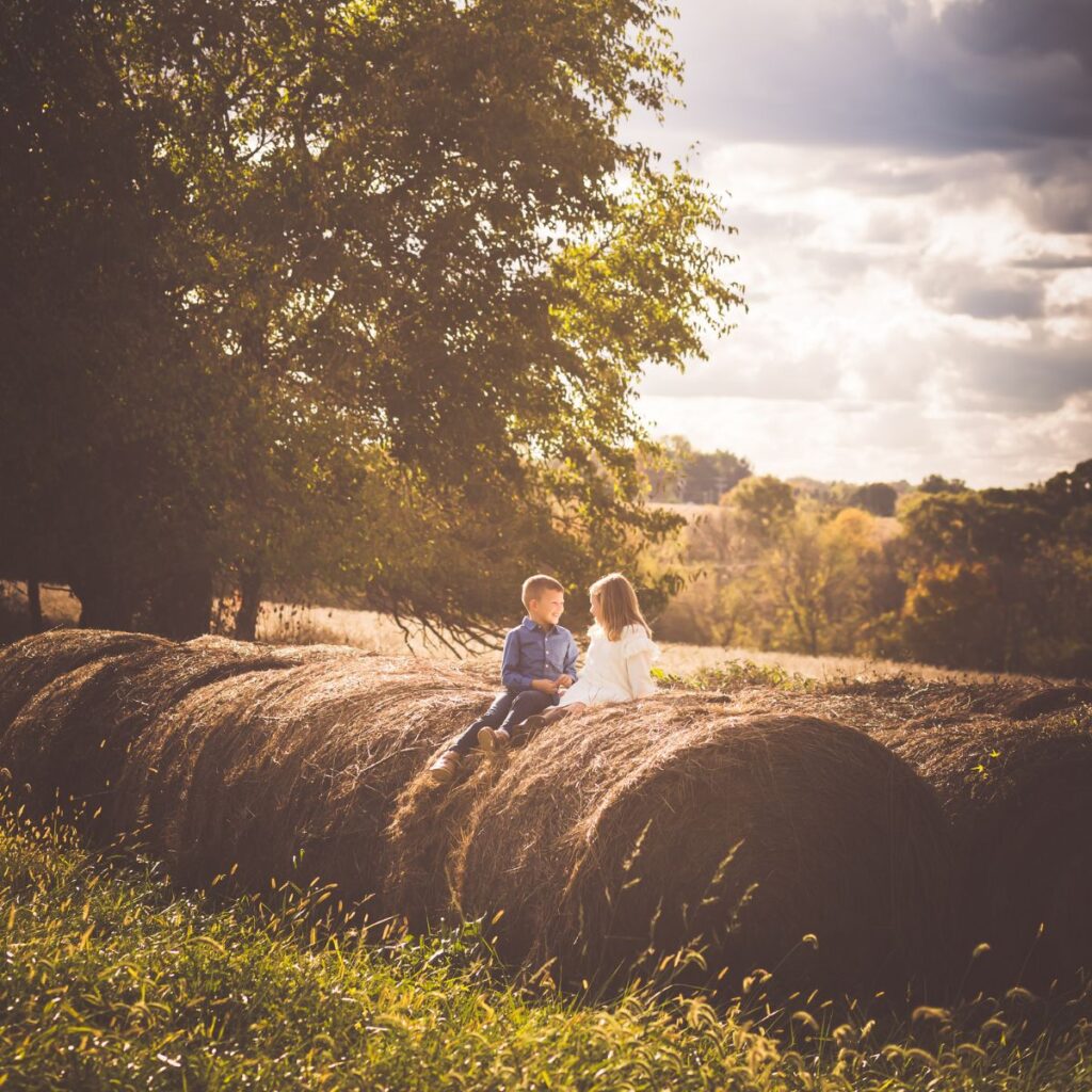 Two children sit on large hay bales in a sunlit, rural field. A tree and cloudy sky form the backdrop, creating a warm, serene atmosphere.