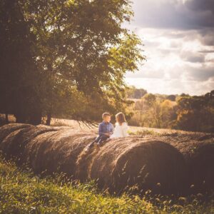 Two children sit on large hay bales in a sunlit, rural field. A tree and cloudy sky form the backdrop, creating a warm, serene atmosphere.
