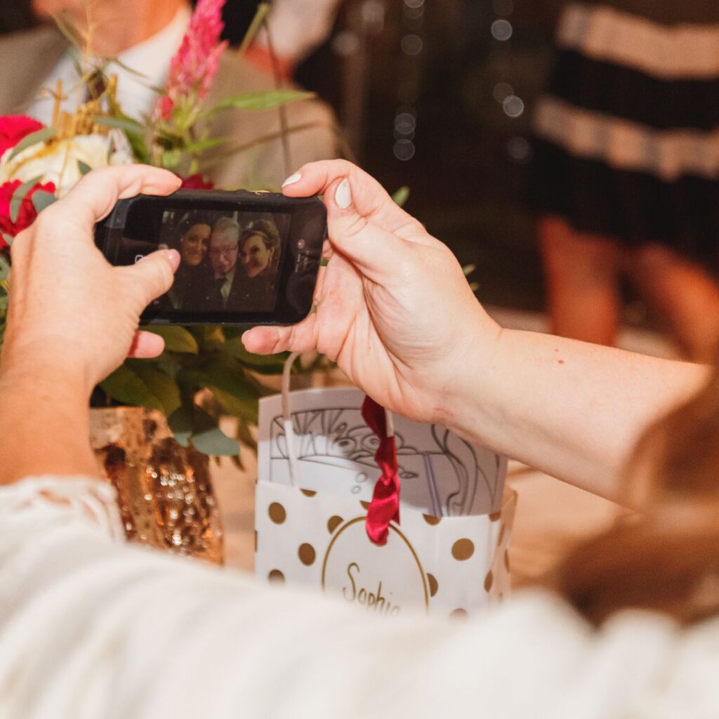 Hands holding a smartphone capturing a group selfie near a floral arrangement. A white gift bag with gold dots and "Sophie" on it is in the foreground.