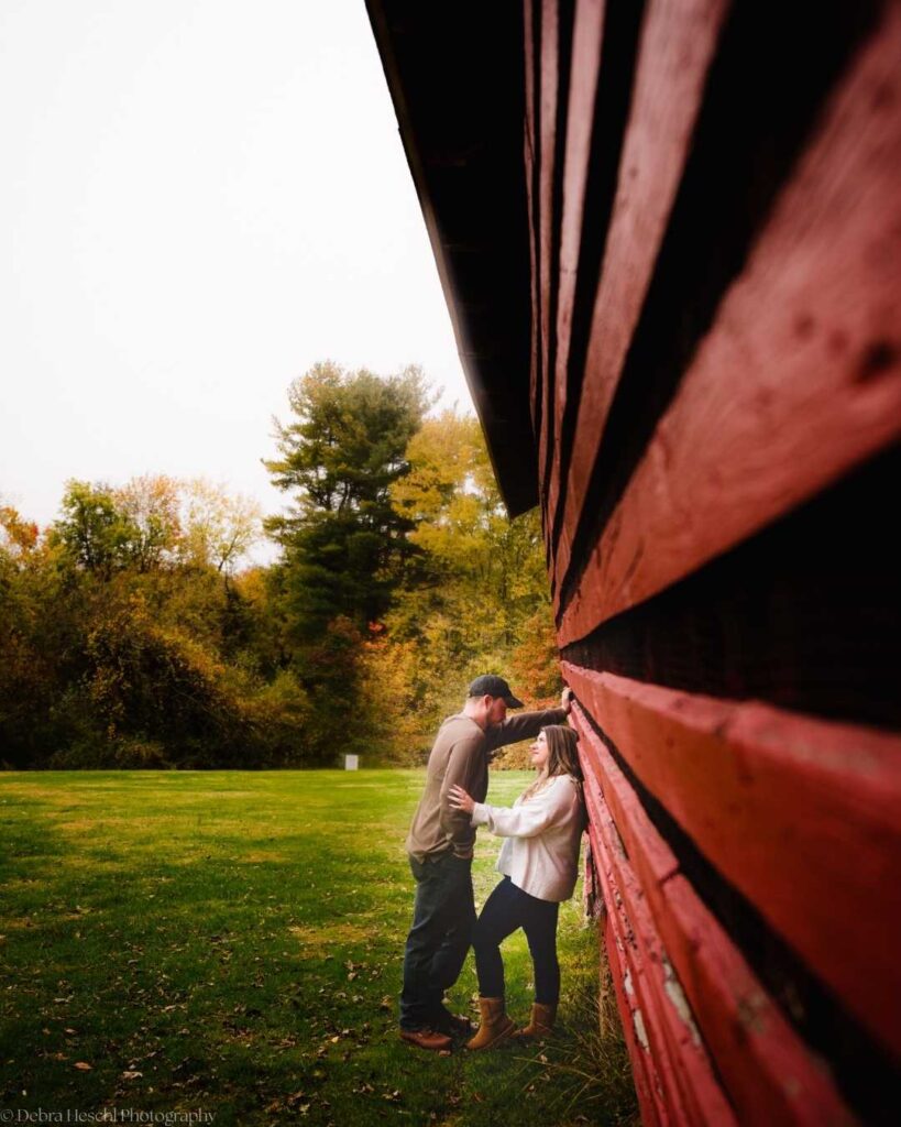 A couple leans against a red barn, sharing an intimate moment. The scene is set in a lush, green field with colorful autumn trees, conveying warmth and romance.